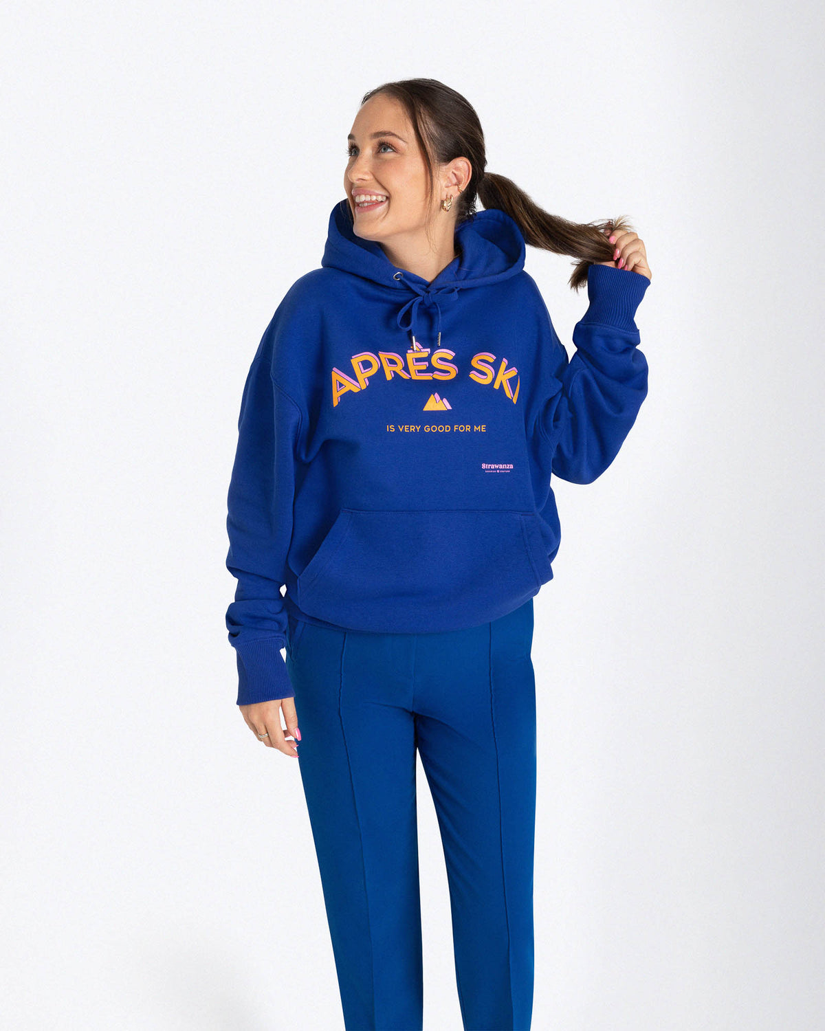 Entdecken Sie den stylischen 'APRES SKI HOODIE' von Strawanza, ideal für Outdoor-Abenteuer. Das Model präsentiert diesen trendigen blauen Hoodie in einer natürlichen Kulisse, die Funktionalität und Modebewusstsein vereint. Der Hoodie zeichnet sich durch sein starkes blau und den Siebdruck im Retro Stil aus. Perfekt für Berg- und Skiabenteuer. Hieran einem Frauenmodel zu sehen.