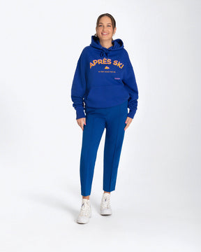 Entdecken Sie den stylischen 'APRES SKI HOODIE' von Strawanza, ideal für Outdoor-Abenteuer. Das Model präsentiert diesen trendigen blauen Hoodie in einer natürlichen Kulisse, die Funktionalität und Modebewusstsein vereint. Der Hoodie zeichnet sich durch sein starkes blau und den Siebdruck im Retro Stil aus. Perfekt für Berg- und Skiabenteuer. Hier an einem Frauenmodel zu sehen. 