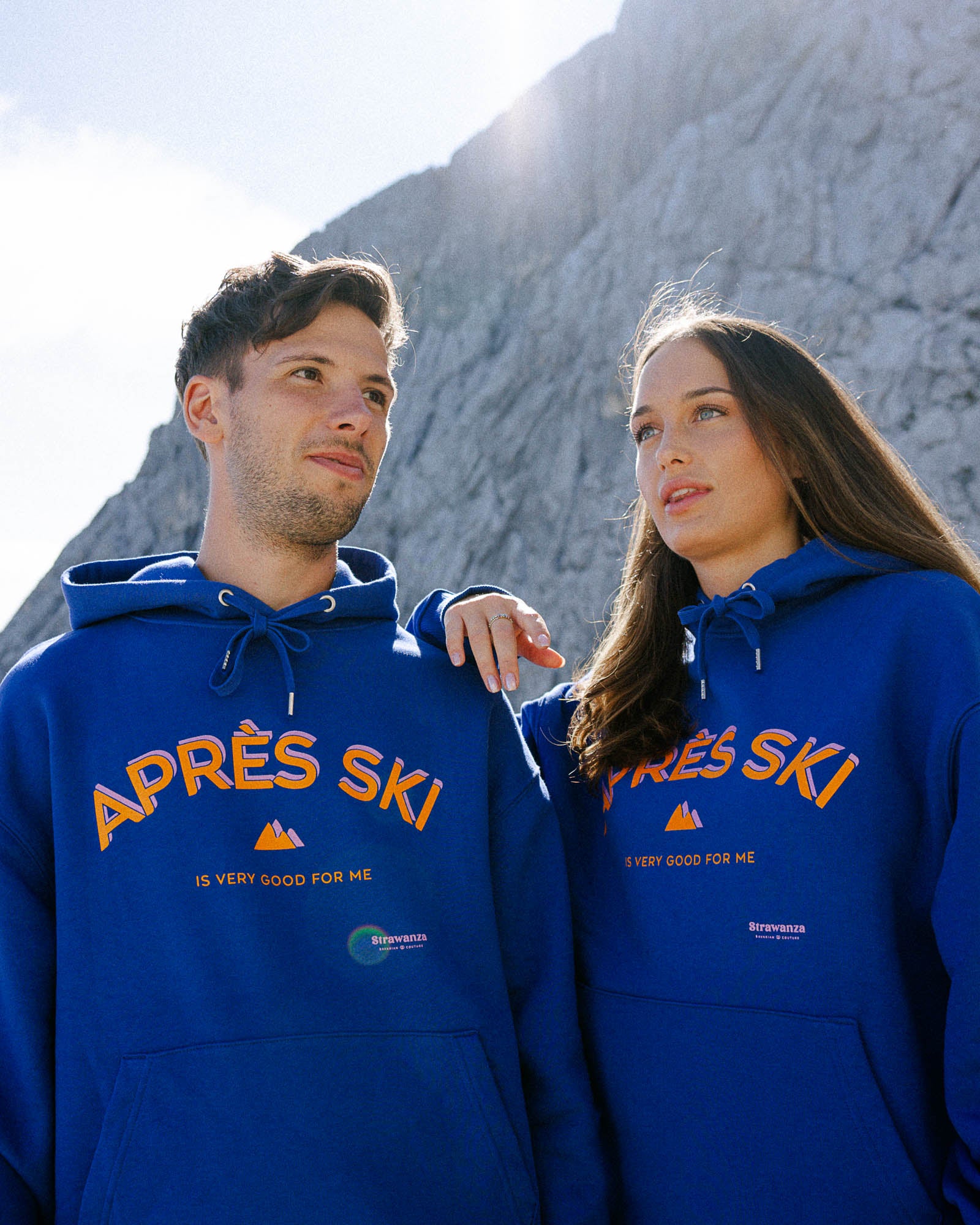 Entdecken Sie den stylischen 'APRES SKI HOODIE' von Strawanza, ideal für Outdoor-Abenteuer. Das Model präsentiert diesen trendigen blauen Hoodie in einer natürlichen Kulisse, die Funktionalität und Modebewusstsein vereint. Der Hoodie zeichnet sich durch sein starkes blau und den Siebdruck im Retro Stil aus. Perfekt für Berg- und Skiabenteuer. Hier an einem Männermodel und einem Frauenmodel zu sehen. 